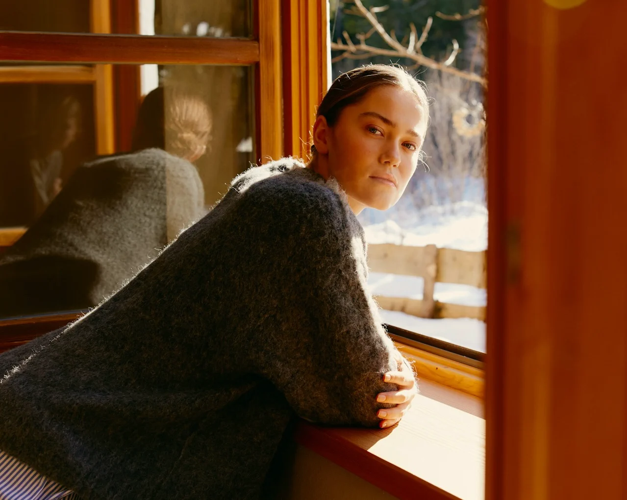 Young woman with braided hair leaning on a windowsill and looking out at a snowy landscape.