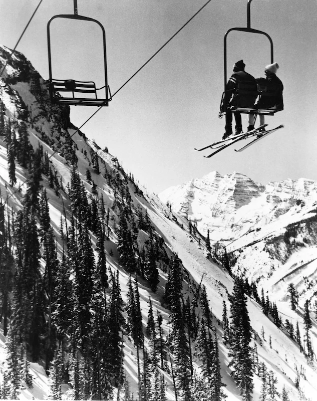 Two skiers ride a ski lift up a snow-covered mountain with evergreen trees.