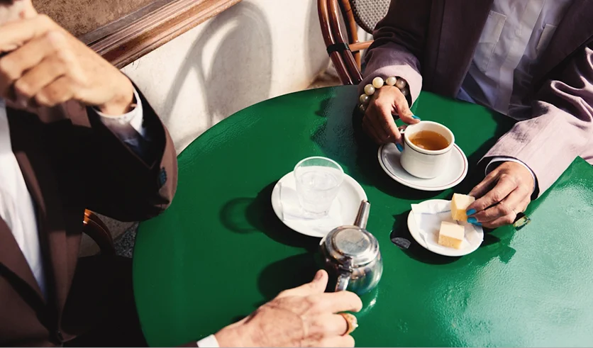 Two people are seated at a green table, enjoying tea and buttered bread.