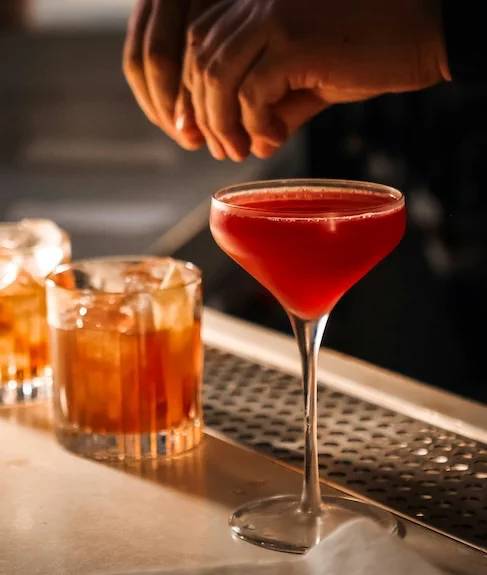Close-up of a bartender garnishing a red cocktail with a lemon peel.
