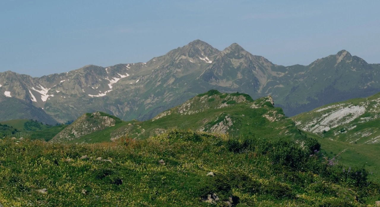 A rugged mountain range under a clear blue sky, with green, rolling hills in the foreground.