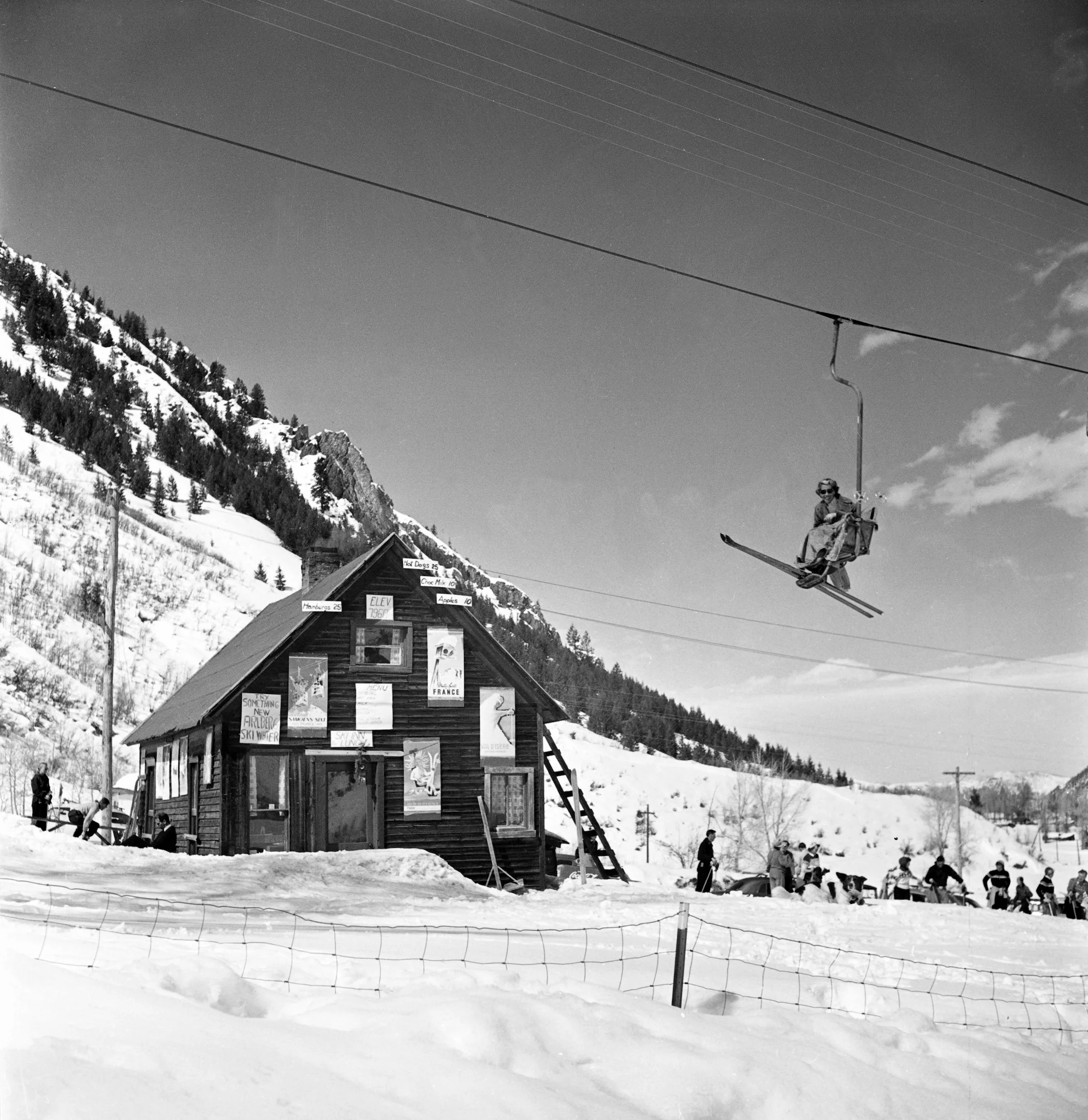 Black and white photo of a skier mid-air above a rustic ski lodge with various posters on its side.