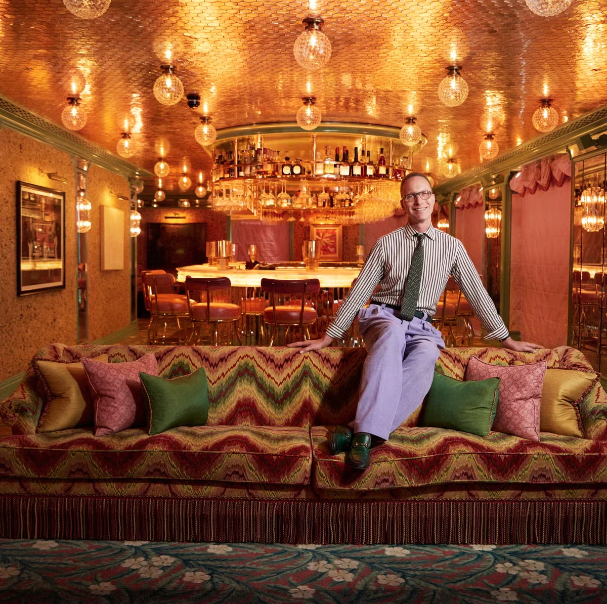 A man in glasses, a green tie, and lavender pants sits on a patterned couch in a richly decorated room with a bar in the background.